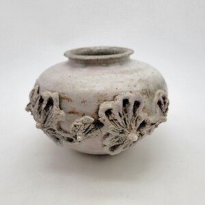 May include: A small, round ceramic vase with a pale, off-white glaze and a textured surface. The exterior features raised, sculpted floral motifs in a muted, earthy tone that wrap around the midsection. The rim is circular and slightly darkened.