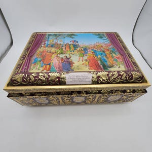 May include: A rectangular metal box featuring a colourful illustration of a crowd scene on the lid, framed by purple curtains and gold embossed floral patterns. The text on the lid reads: Die Meistersinger von Nürnberg Oper von Richard Wagner.