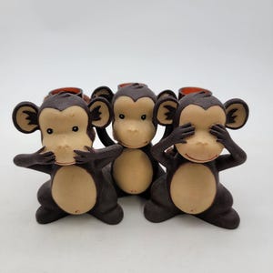 Può includere: Tre statuette di scimmia nella classica posa "Non vedere il male, non sentire il male, non dire il male". Ogni scimmia ha un viso marrone chiaro e un corpo marrone scuro, con le mani che coprono gli occhi, le orecchie e la bocca.