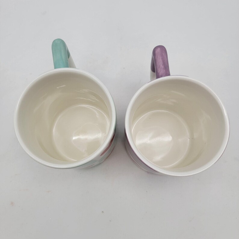 May include: Two white ceramic mugs with pastel-colored handles. One mug has a light green handle, and the other has a purple handle. The mugs are empty and have a glossy finish.