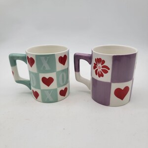 May include: Two ceramic mugs. One mug has a mint green handle and a checkered pattern with red hearts and the letters X and O. The other mug has a purple handle and a checkered pattern with a red flower and red hearts.