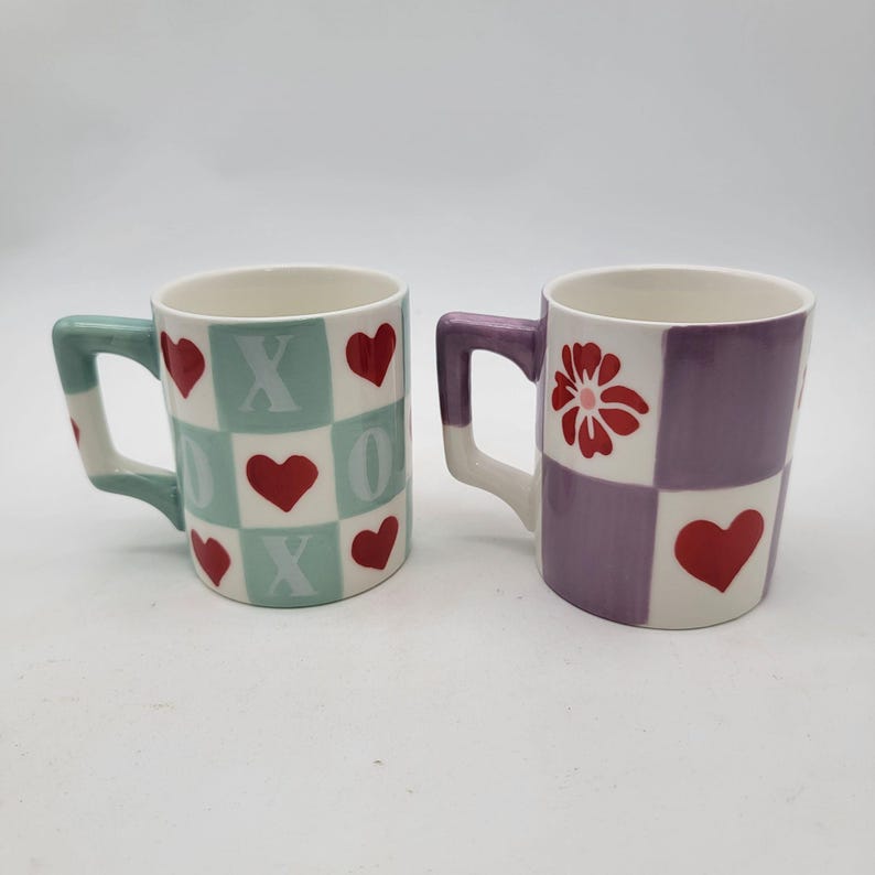 May include: Two ceramic mugs. One mug has a mint green handle and a checkered pattern with red hearts and the letters X and O. The other mug has a purple handle and a checkered pattern with a red flower and red hearts.