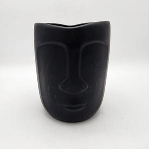 May include: A matte black ceramic vase with a face design. The vase has a smooth, rounded shape with a carved face featuring a nose, eyes, and a subtle smile. The vase is a decorative item.
