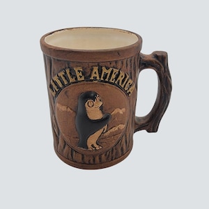 Könnte beinhalten: Braune Keramiktasse mit Baumrindenstruktur und einem astförmigen Griff. Die Tasse zeigt ein rundes Emblem mit einer Pinguin-Illustration und den Worten "LITTLE AMERICA" in goldfarbenen Buchstaben. Der Innenraum ist cremefarben.