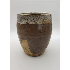May include: A handmade ceramic tumbler with a rustic design. The cup features a brown body with a lighter brown and white rim. The bottom has a lighter tan color. The tumbler is suitable for hot or cold beverages.