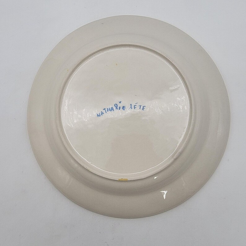 Puede incluir: Un plato de cer&aacute;mica de color crema con interior blanco. La parte inferior del plato presenta el texto manuscrito "Nathalie Lete" en azul. El plato es redondo y parece hecho a mano.