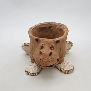 Peut inclure: Un pot de fleurs en céramique marron en forme de tête d'hippopotame avec une ouverture large sur le dessus. L'hippopotame a de petits yeux sombres, un museau texturé avec des narines pointillées et porte des chaussures blanches aux pattes avant.