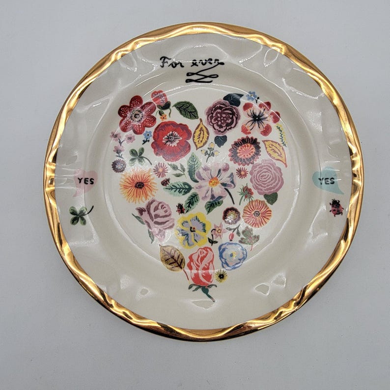 Puede incluir: Cenicero color crema con borde dorado. El centro presenta un arreglo floral en forma de coraz&oacute;n en varios colores. Las palabras "For ever" y "YES" est&aacute;n escritas en el cenicero.