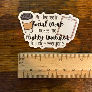 以下が含まれることがあります： コーヒーカップとクリップボードが描かれた白いステッカー。テキストは「ソーシャルワークの学位は、私を非常に有資格にし、すべての人を判断することができます。」