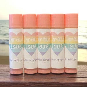 May include: Five lip balm tubes with peach-colored caps and labels. The labels feature a rainbow heart design with the words "Thread Love" and "Ocean Breeze". The tubes are arranged on a wooden surface with a blurred ocean background.