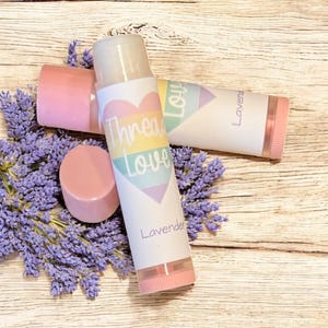 May include: Two lavender-scented lip balm tubes with pink and white packaging, one with a matching pink cap. The tubes are decorated with a heart-shaped design and the words "Thread Love". The lip balms are surrounded by sprigs of lavender.