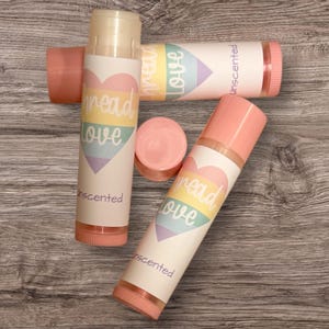May include: Lip balm tubes with pink caps and bases, and white labels. The labels feature a rainbow heart design with the words "spread love" and "unscented". The lip balms are arranged on a wooden surface.