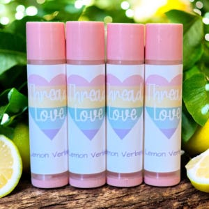 May include: Four lip balm tubes with pink caps and labels featuring a pastel heart design and the words "Thread Love." The bottom of the labels read "Lemon Verbena." The background includes green foliage and lemons.
