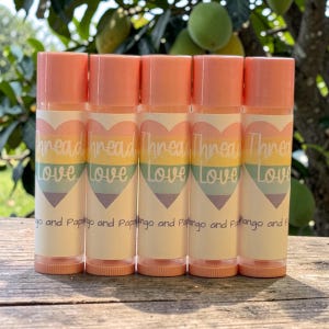 May include: Five lip balm tubes with peach-colored caps and bases. Each tube features a rainbow heart design with the words "Thread Love" and additional text. The lip balms are arranged on a wooden surface.