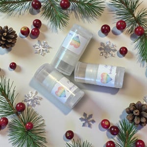 May include: Three clear lip balm tubes with white labels featuring a rainbow heart design and the words "Pure Love" and "babygirl". The tubes are surrounded by pine needles, red berries, silver snowflakes, and pine cones, creating a festive holiday theme.