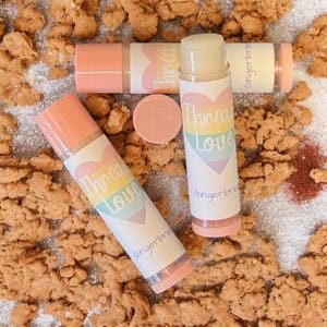 May include: Three lip balms with pink and white labels are displayed on a bed of cookie crumbs. The labels feature a rainbow heart design with the words "Thread Love" and "Gingerbre". One lip balm has its cap off, revealing the product.