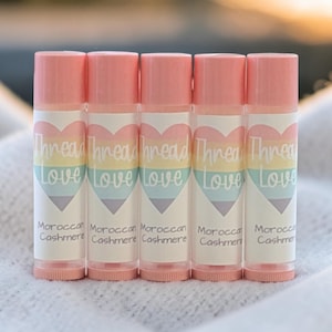 May include: Five lip balm tubes with pink caps and labels. The labels feature a rainbow heart design with the words "Thread Love" and "Moroccan Cashmere". The tubes are arranged in a row.