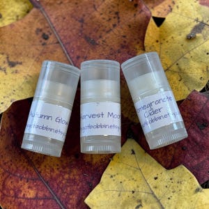 May include: Three clear plastic lip balm tubes with white labels. The labels feature handwritten text reading Autumn Glow, Harvest Moon, and Pomegranate Cider, each followed by the web address gotbobbin.etsy.com in black ink.