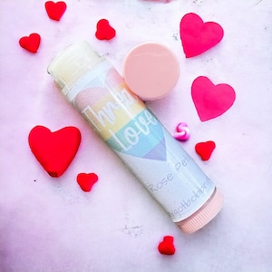 May include: A pink lip balm tube with a rainbow heart design and the text "Thrive Love" on the front. The tube also has the text "Rose Petals" and "Naturally Delicious" on the side. The lip balm is surrounded by red hearts.