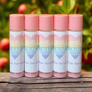 May include: Five lip balm tubes with pink caps and labels featuring a rainbow heart design. The labels read "Thread Love" and "Apple Harvest". The tubes are arranged vertically against a blurred background.