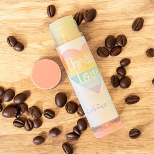 May include: A rainbow heart with the text "Throm Love" and "Coffee Co." on a white and pink lip balm tube. The tube is surrounded by coffee beans on a wooden surface.