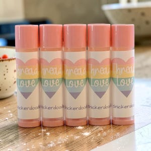 May include: Five cylindrical lip balm tubes with light pink caps. Each white label features a pastel rainbow heart graphic with the text Thread Love in white script and the word snickerdoodle printed in small black letters at the bottom.