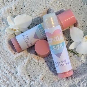 May include: Two cylindrical lip balm tubes with pink caps and white labels. The labels feature a pastel rainbow heart graphic with the text "Thread Love" and "Sea Salt and Orchid" printed in dark blue. Two white orchid flowers are placed nearby.