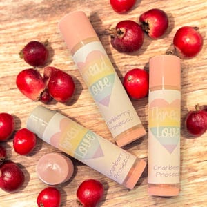May include: Three pink lip balm tubes with rainbow heart designs and the text "Thread Love" on the labels. The lip balms are on a wooden surface with red berries scattered around them. The lip balms are labeled "Cranberry Prosecco".