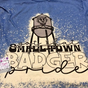 May include: A blue t-shirt with a white bleach design. The design features a water tower with a badger on it and the text "got that small town badger pride".
