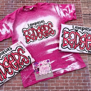 May include: Three pink and white bleached t-shirts with the text "Lampasas Badgers" in red and black lettering. The shirts are on a brick background.