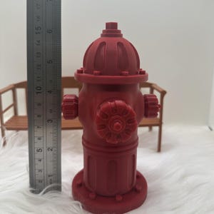 May include: A red fire hydrant toy with a detailed design. The hydrant is about 6 inches tall and has a round top with a small knob on top. The hydrant has a black handle on each side and a black cap on the front.