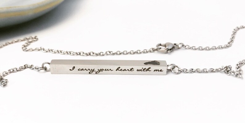 May include: A silver necklace with a bar pendant engraved with the words "I carry your heart with me" and a small footprint. The necklace is on a white background.