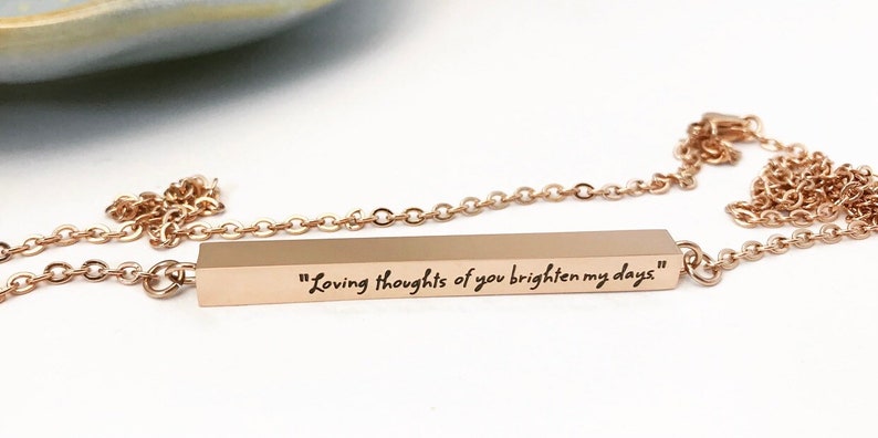 May include: A rose gold bar necklace with a chain and a bar pendant engraved with the text "Loving thoughts of you brighten my days."