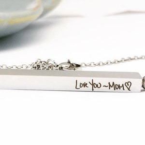 May include: A silver bar necklace with a chain. The bar has the words "Love You - Mom" engraved on it.