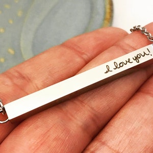 May include: A silver bar necklace with a chain. The bar is engraved with the words "I love you!" in cursive handwriting.