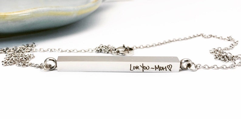 May include: A silver necklace with a bar pendant engraved with the words "Love You - Mom" and a heart symbol.