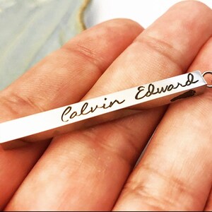May include: A silver bar necklace with a delicate chain. The bar is engraved with the name "Calvin Edward" in cursive script.