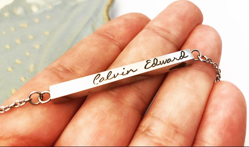 May include: A silver bar necklace with a delicate chain. The bar is engraved with the name "Calvin Edward" in cursive script.