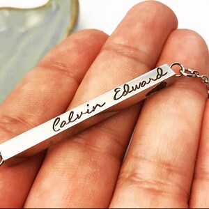 May include: A silver bar necklace with the names "Calvin Edward" engraved on it. The bar has a small heart shape at the end.