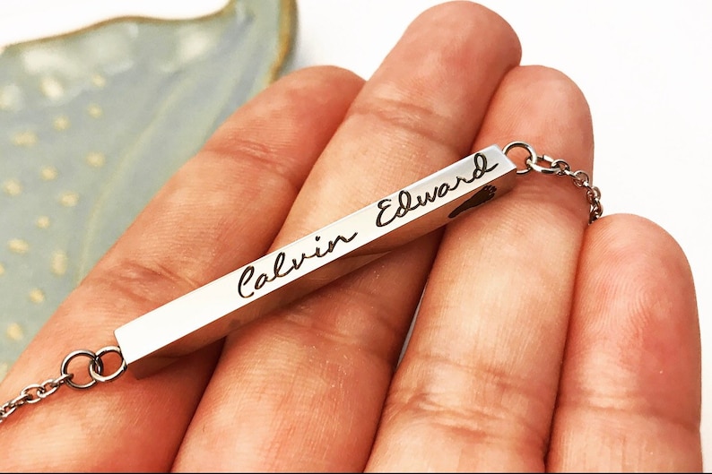 May include: A silver bar necklace with the names "Calvin Edward" engraved on it. The bar has a small heart shape at the end.