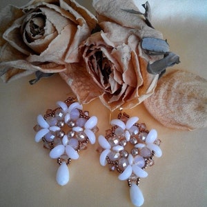 Pode incluir: Um par de brincos de contas brancas e marrons com um pingente em forma de lágrima. Os brincos são feitos com pequenas contas brancas e contas de sementes marrons.