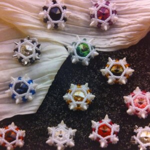 May include: A collection of beaded pendants with various colored crystal centers, including black, green, blue, and red. Each pendant has a spiky, star-like design with white and silver beads. The pendants are displayed on a textured surface.
