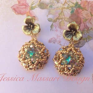 Pode incluir: Brincos dourados com uma parte superior em forma de flor e uma gota esférica com contas e cristais iridescentes. Os brincos estão sobre um fundo floral suave. Texto: Jessica Massari Design.