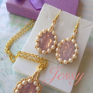 Pode incluir: Um conjunto de joias delicadas com um pingente e brincos com um cristal rosa cercado por pérolas brancas e detalhes dourados. O conjunto é apresentado em uma superfície branca com a palavra "Jessy" escrita em rosa.