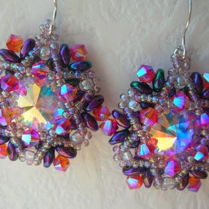 Pode incluir: Um par de brincos com um centro de cristal roxo, rosa e iridescente rodeado por contas iridescentes e fio de prata.