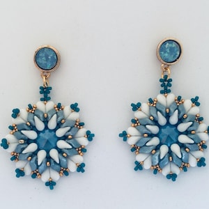 Puede incluir: Un par de pendientes hechos a mano con un diseño de copo de nieve. Los pendientes presentan una gema azul central rodeada de cuentas blancas y azules en forma de lágrima. Acentos dorados y cuentas turquesas añaden detalles.
