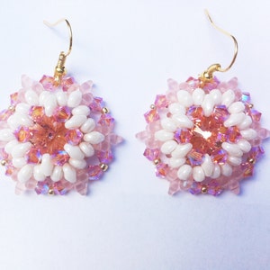 Pode incluir: Um par de brincos redondos de contas rosa e brancas com ganchos dourados. Os brincos têm um design floral.