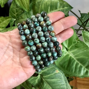 May include: A strand of turquoise beads, with a mottled green and brown pattern, is wrapped around a hand. The beads are round and have a smooth, polished finish.