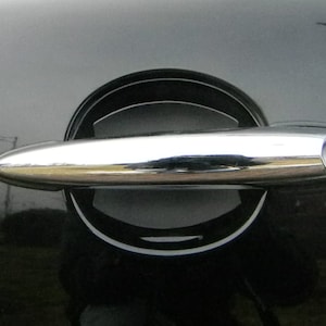 May include: A chrome car door handle with a keyhole. The handle is attached to a black car door.
