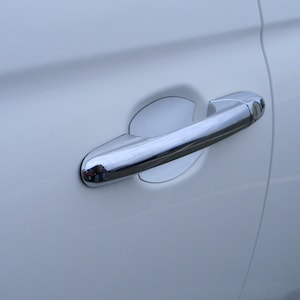 Könnte beinhalten: Eine weiße Autotür mit einem verchromten Türgriff. Der Griff hat eine weiße Kunststoffabdeckung um das Schlüsselloch.